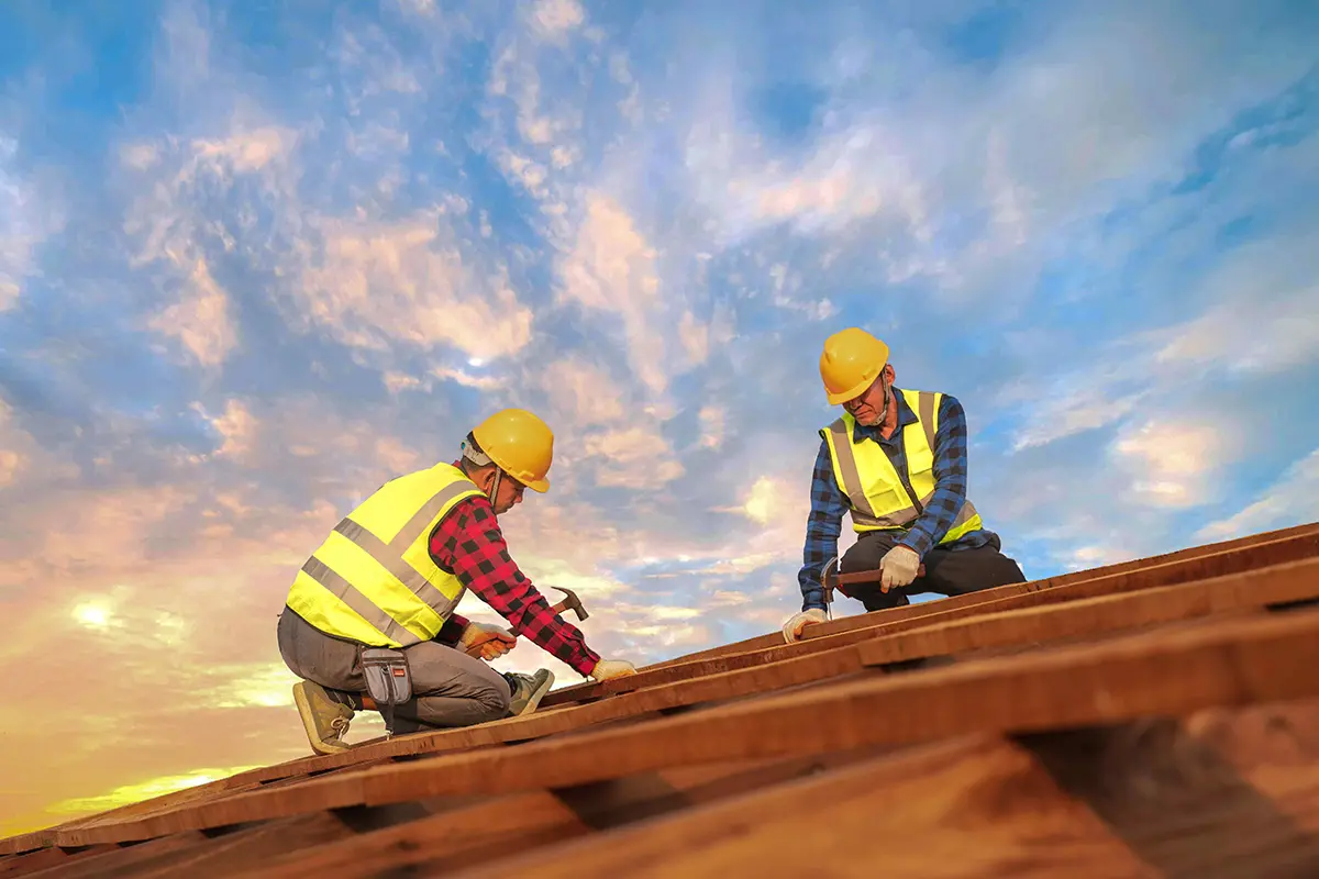 Two carpenters waring high-viz vests and hard hats working on a roof under blue sky.
